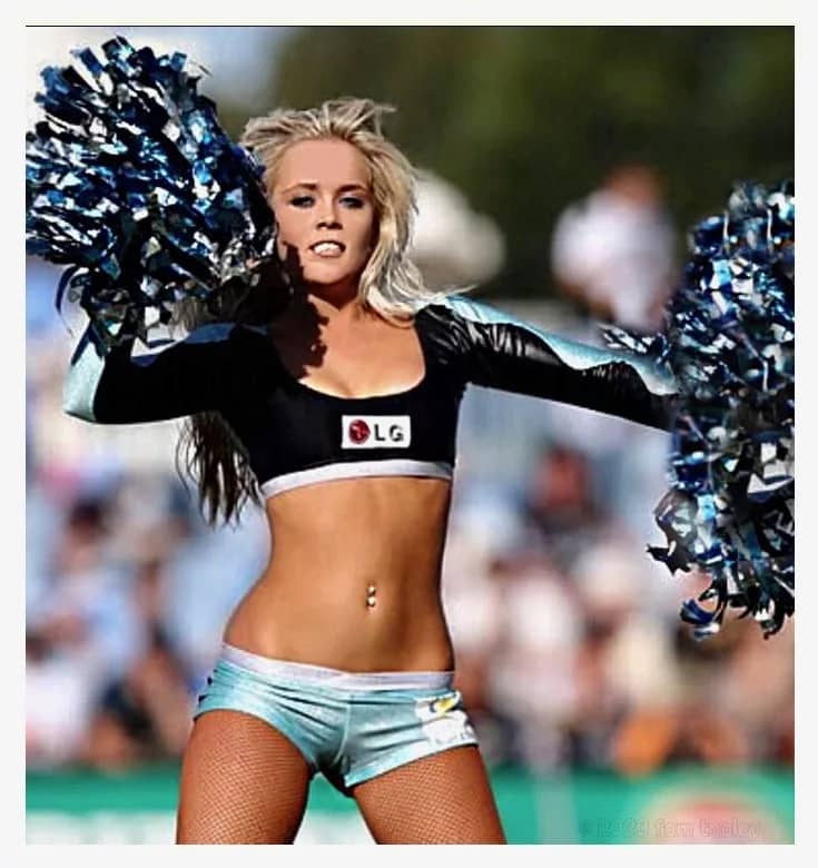 An attractive cheerleader captured via photograph mid-cheer. She is a blinde with a very attractive body - especially because her cheerleader uniform covers so little of her body. She is basically wearing a bikini with sleeves. Her bare midfriff is toned and tanned, and matches her fit legs. In each hand she waves a very large pom pom. She has her belly button pierced.